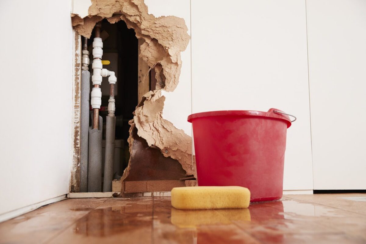 Damaged wall with exposed burst water pipes, sponge, and bucket—evidence of a serious plumbing leak in progress.